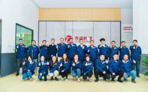 Team photo of Taicheng Machinery staff in blue uniforms giving thumbs-up in front of company logo wall.